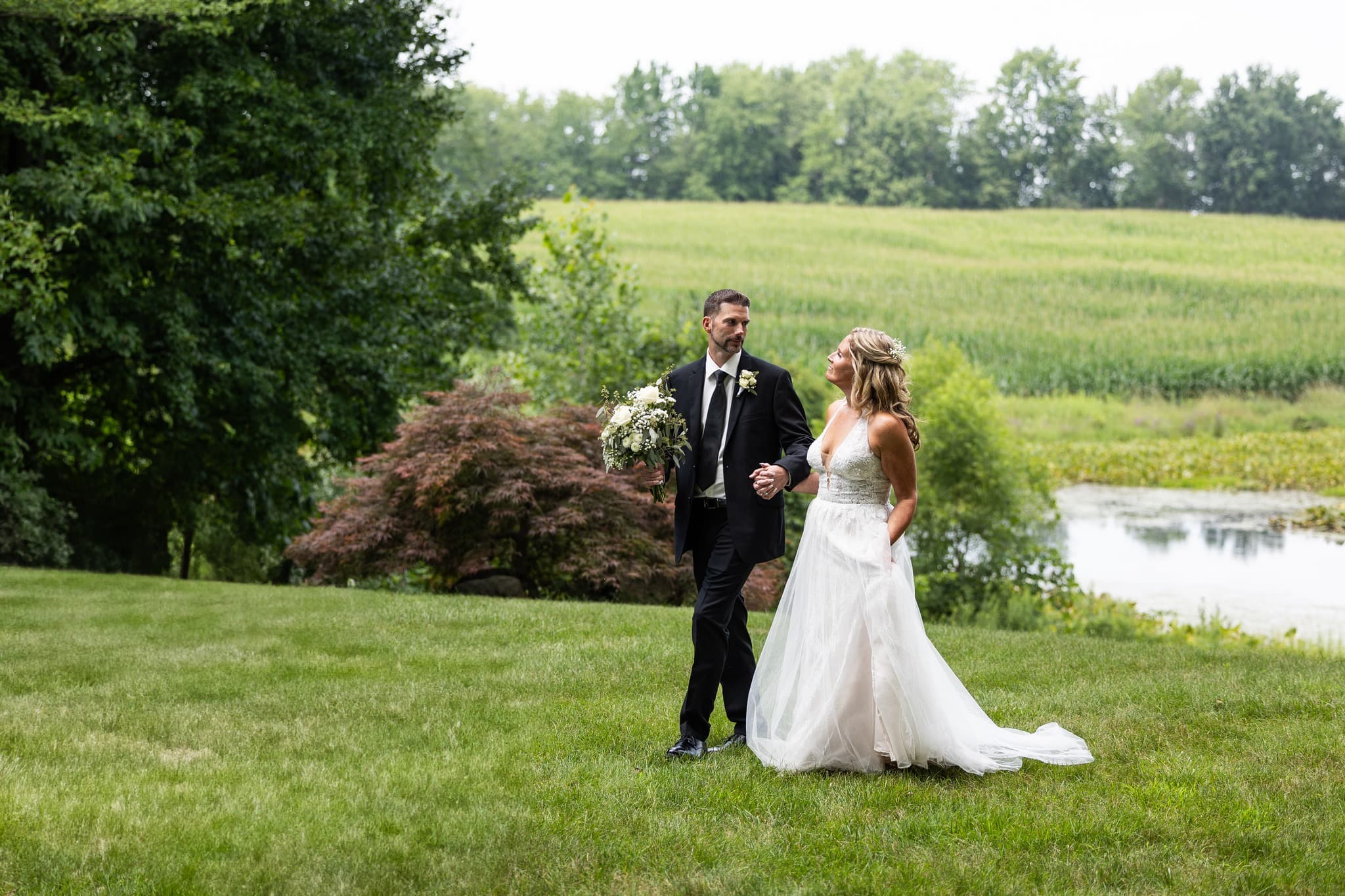 A bride and groom walk hand in hand through a grassy field with lush greenery and a pond in the background.