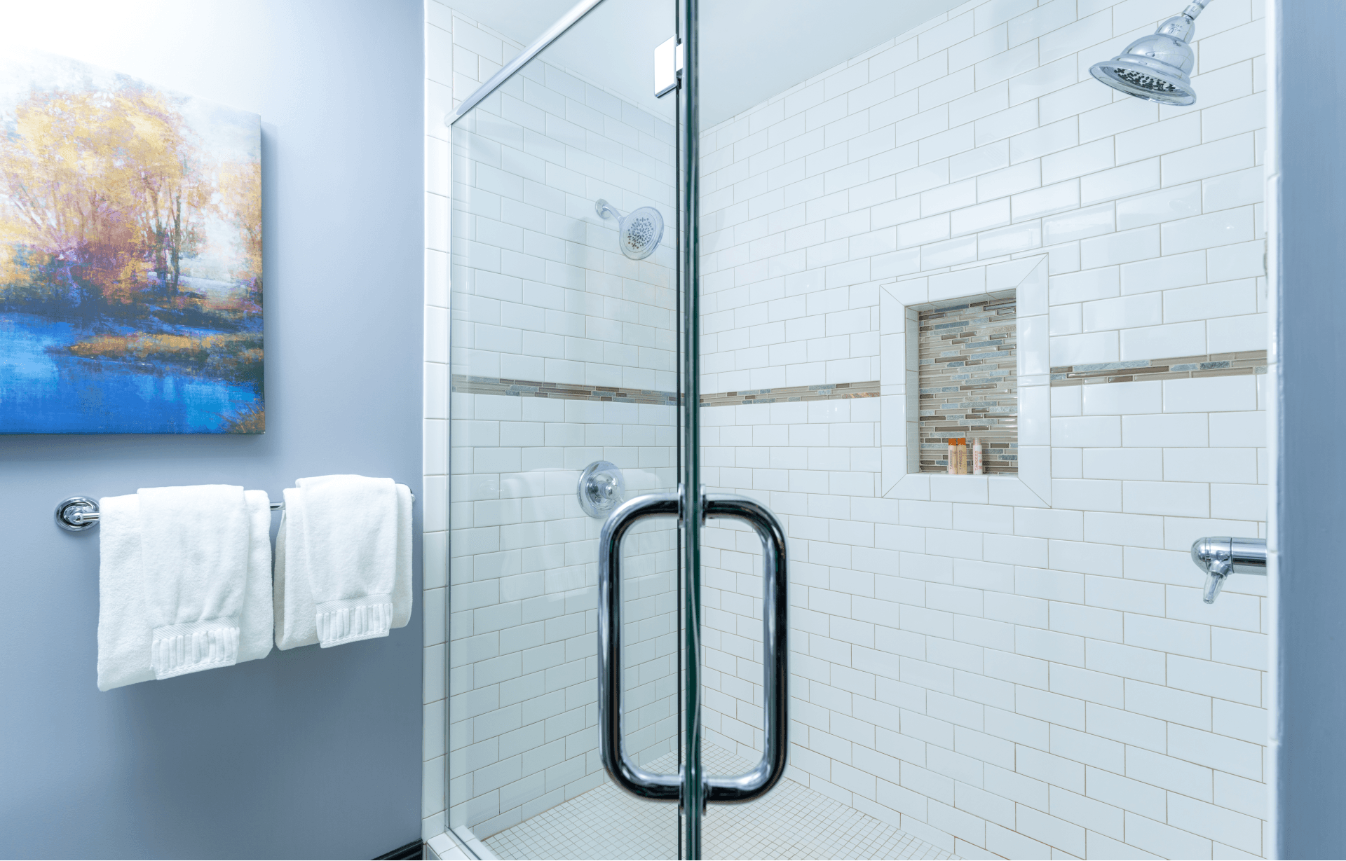 A glass-enclosed walk-in shower with dual shower heads, white subway tile walls, and a decorative niche, next to light blue walls and a scenic painting.