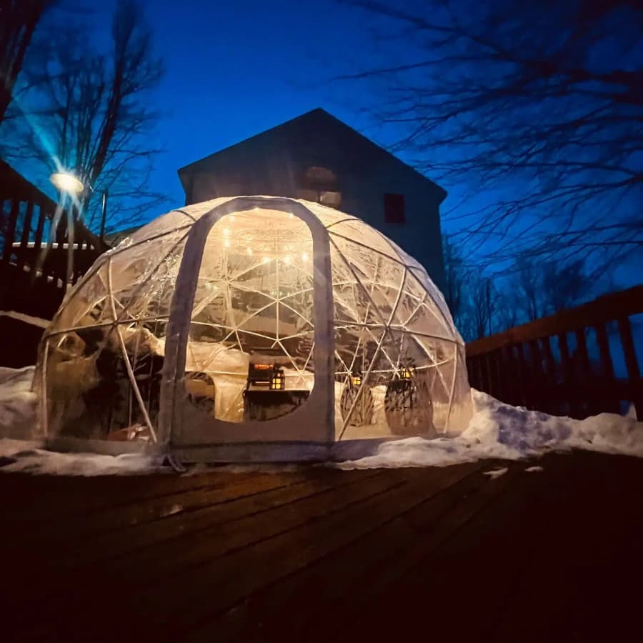 A glowing geodesic dome sits on a snowy deck at dusk, with a house in the background.