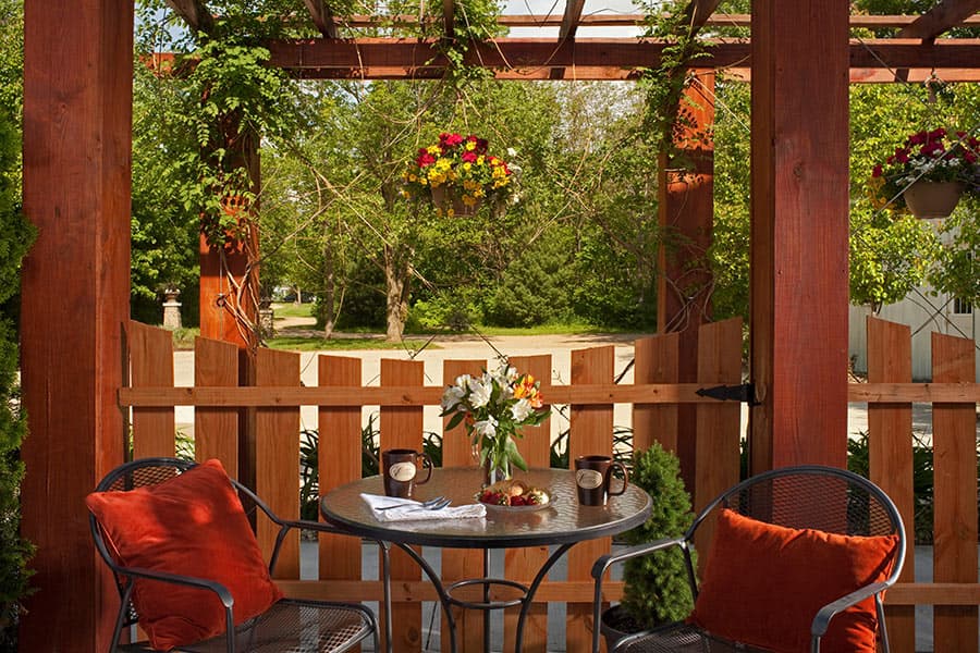 A cozy patio with a small table set for two, surrounded by lush greenery and hanging flower pots.