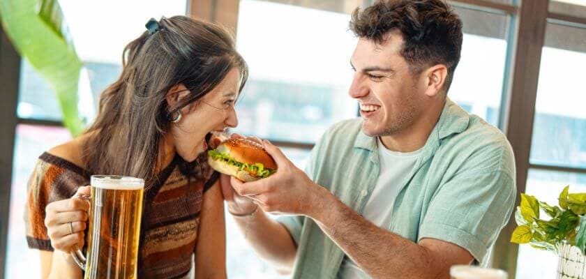 A couple playfully shares a burger while enjoying drinks at a bright indoor setting.