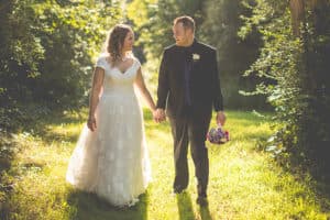 A bride and groom hold hands while walking through a sunlit path in a lush green forest.