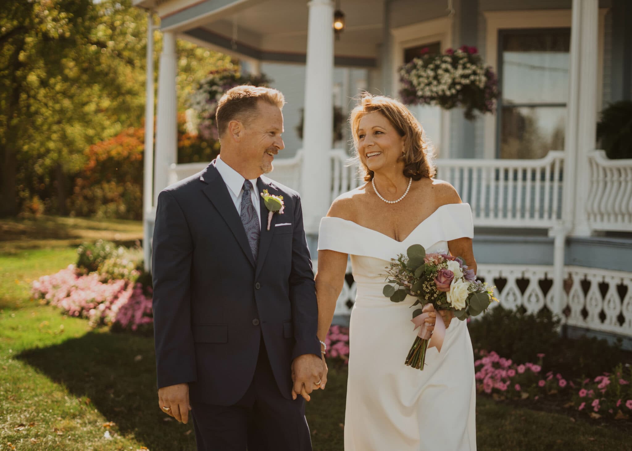 A smiling couple walks hand-in-hand outside a house surrounded by flowers.