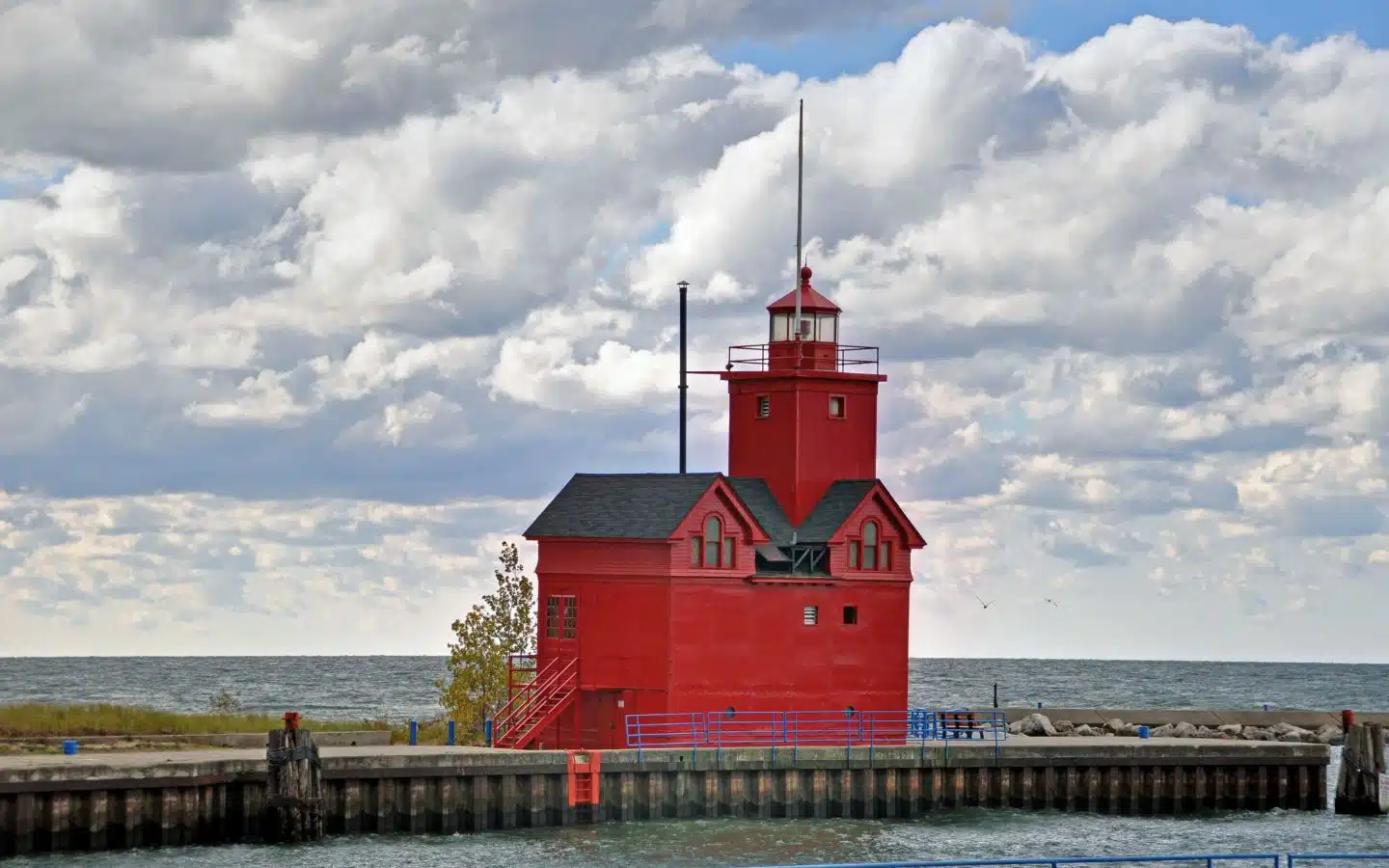 A red lighthouse stands near a pier against a backdrop of cloudy skies and calm waters.