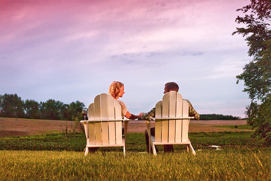 A couple sits hand-in-hand in white chairs, overlooking a serene landscape at sunset.