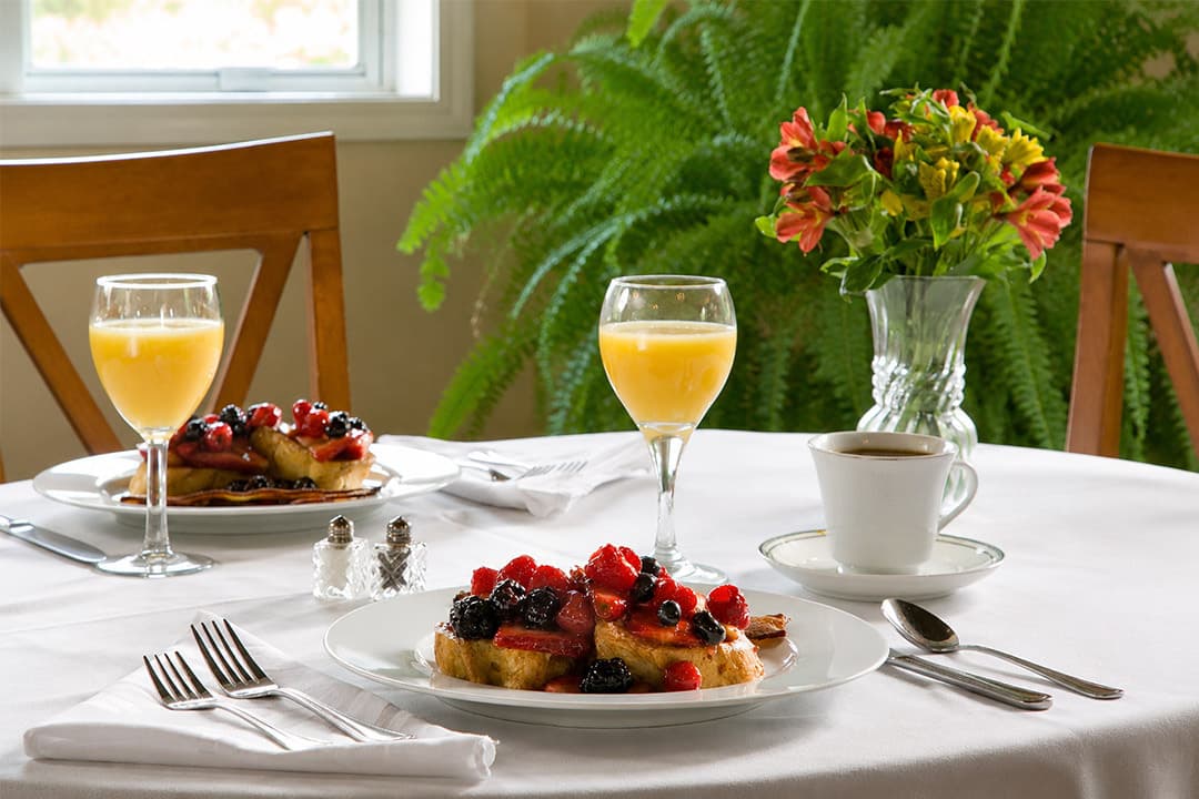 A table set for breakfast featuring French toast topped with berries, two glasses of orange juice, a cup of coffee, and a vase of flowers.