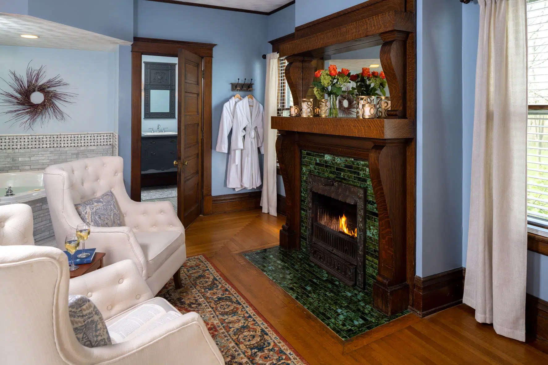 A cozy living room featuring a green-tiled fireplace, two white armchairs, and floral decorations on the mantel.