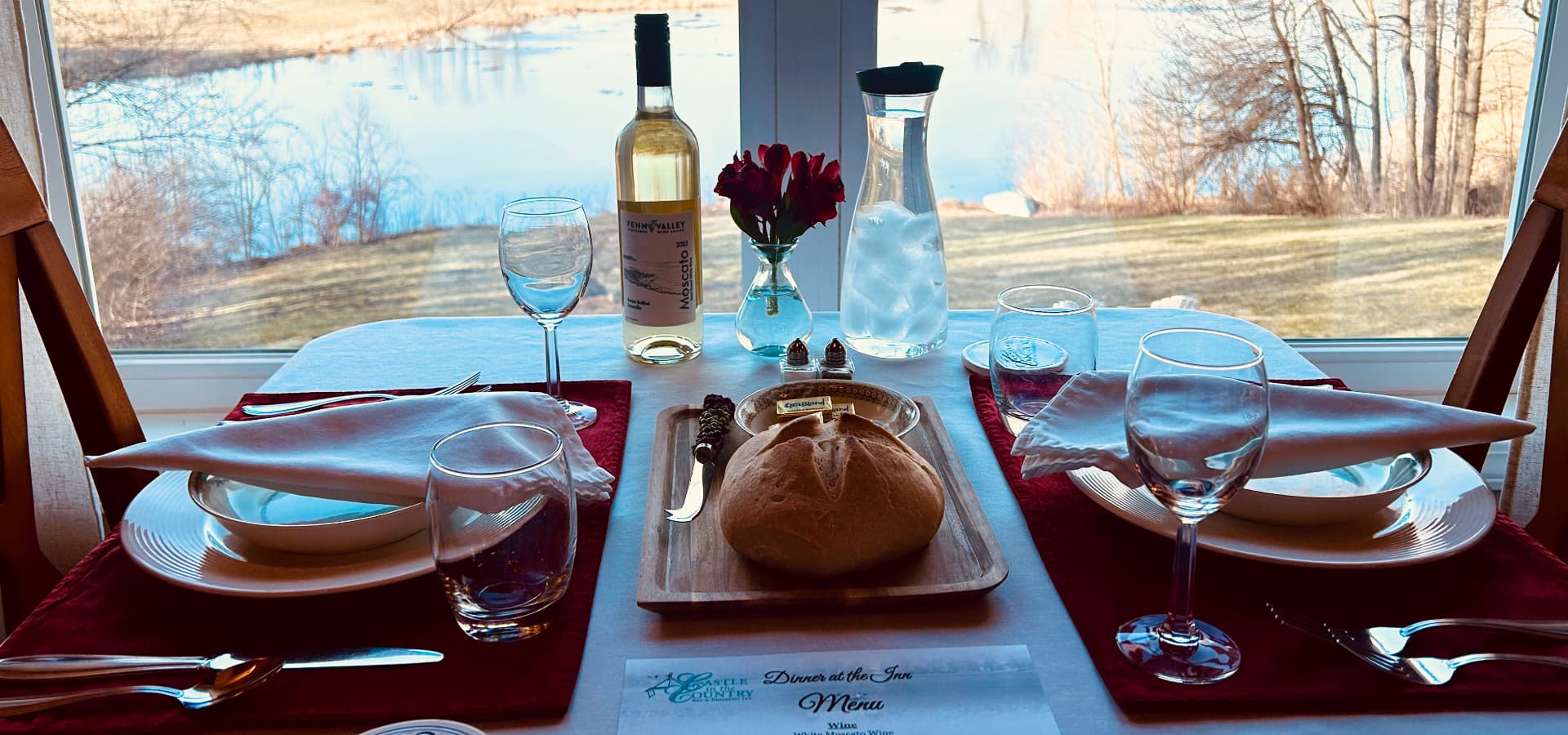 A beautifully set dining table overlooking a scenic lake, featuring wine, glassware, and a loaf of bread.