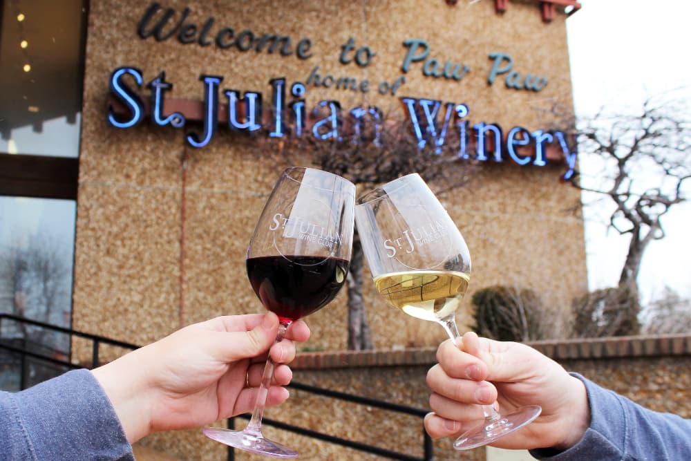Two glasses of wine are clinked in front of the St. Julian Winery sign in Paw Paw.