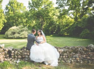 A bride and groom sit closely together on a stone wall surrounded by lush greenery.