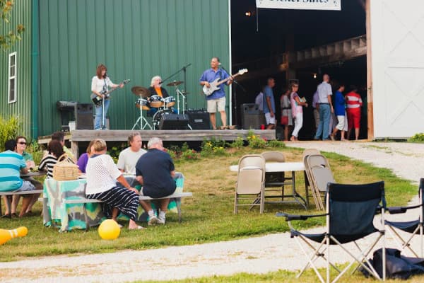 A band performs on a stage while people socialize at picnic tables outside a barn.