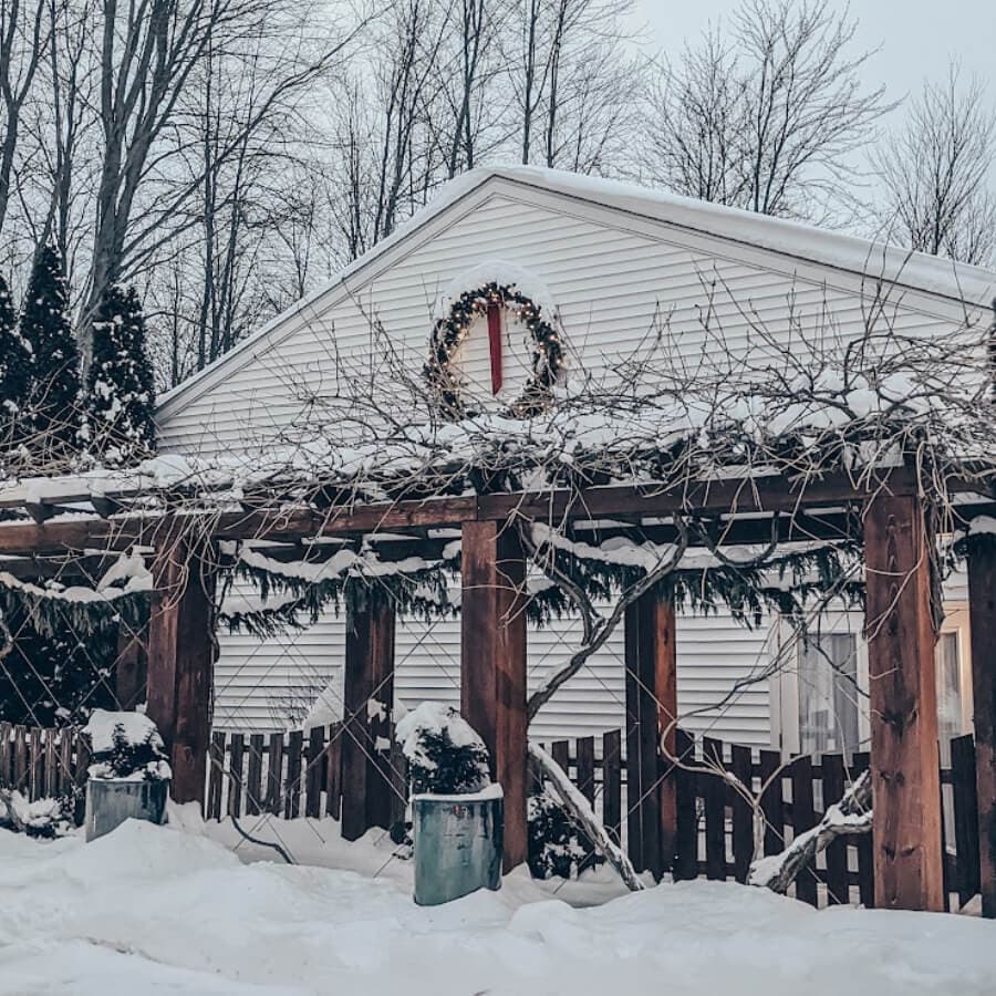 A snowy scene featuring a house adorned with a wreath and a wooden pergola.