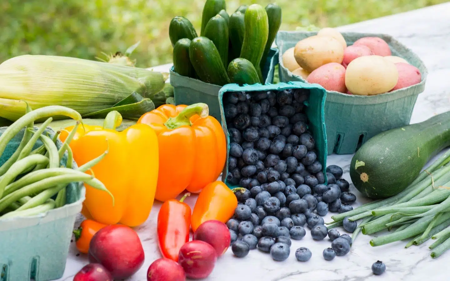 A variety of fresh vegetables and fruits, including peppers, zucchinis, potatoes, corn, green beans, plums, and blueberries, displayed on a table.
