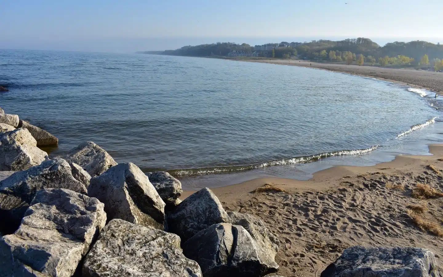 A peaceful beach scene with smooth water, rocks, and distant trees.