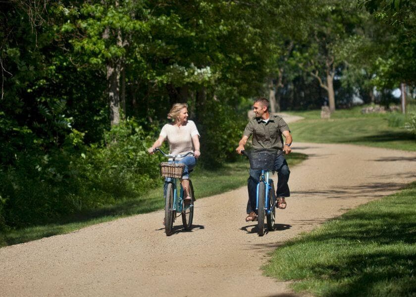 Two people riding bicycles on a gravel path surrounded by greenery.