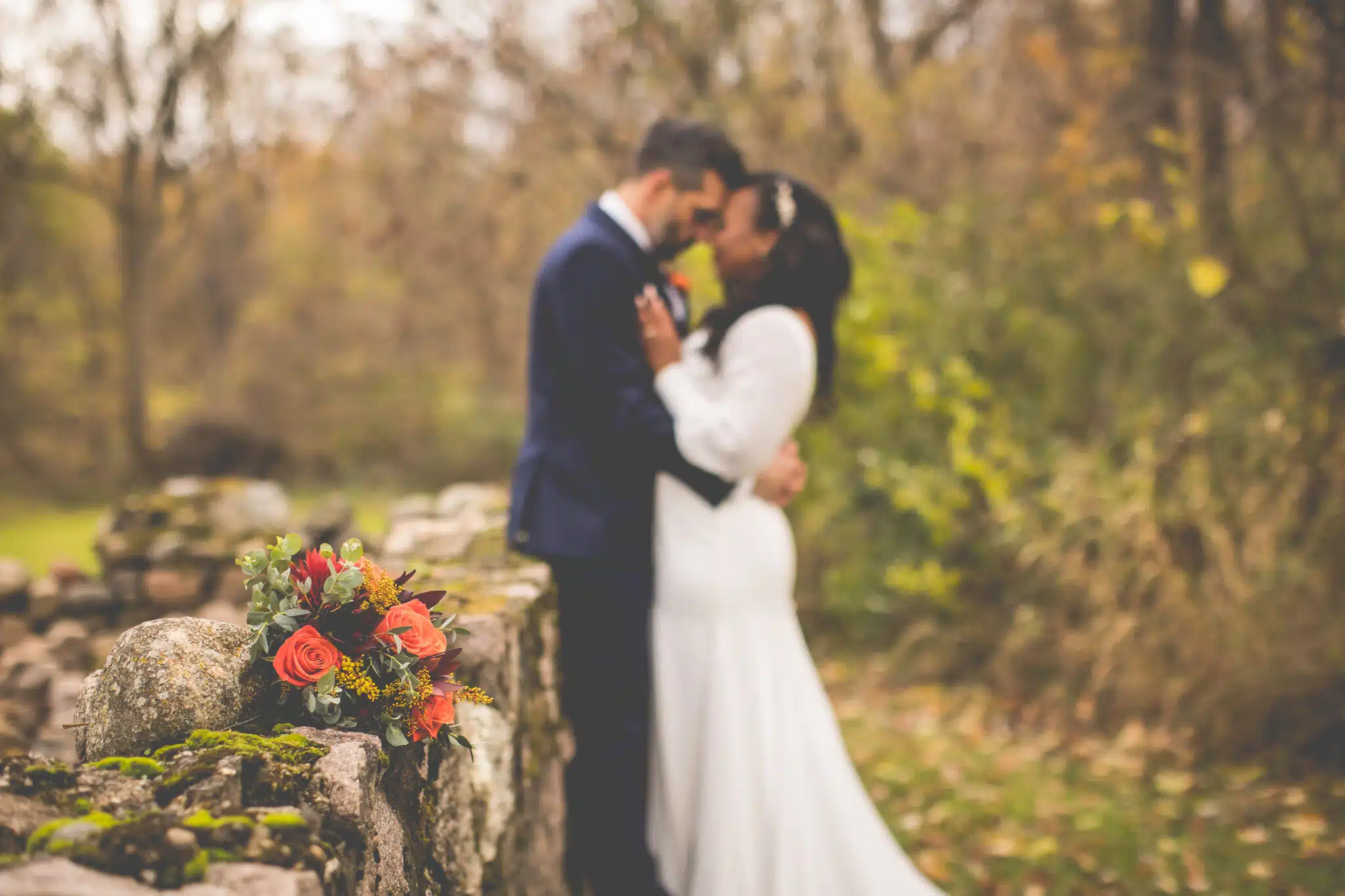 A couple embraces lovingly outdoors, with a bouquet of flowers resting on a stone wall nearby.