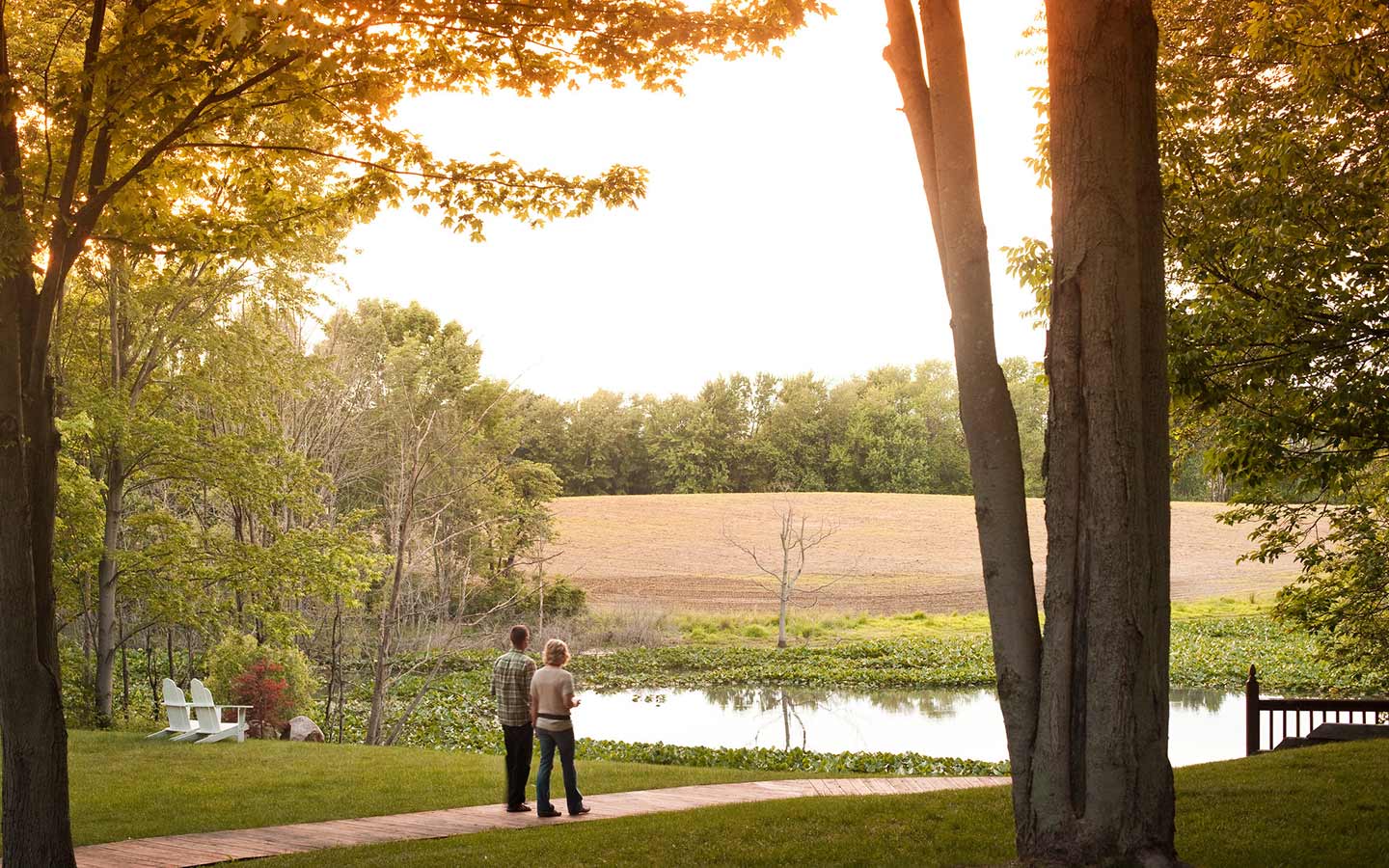 Two people stand by a calm pond at sunset, surrounded by lush greenery and fields.