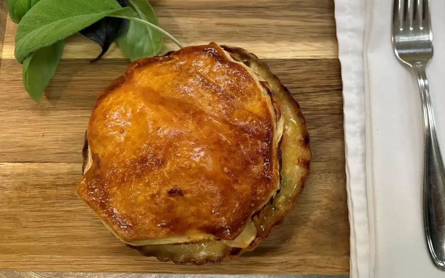 A golden-brown savory pie on a wooden cutting board, accompanied by fresh green leaves.