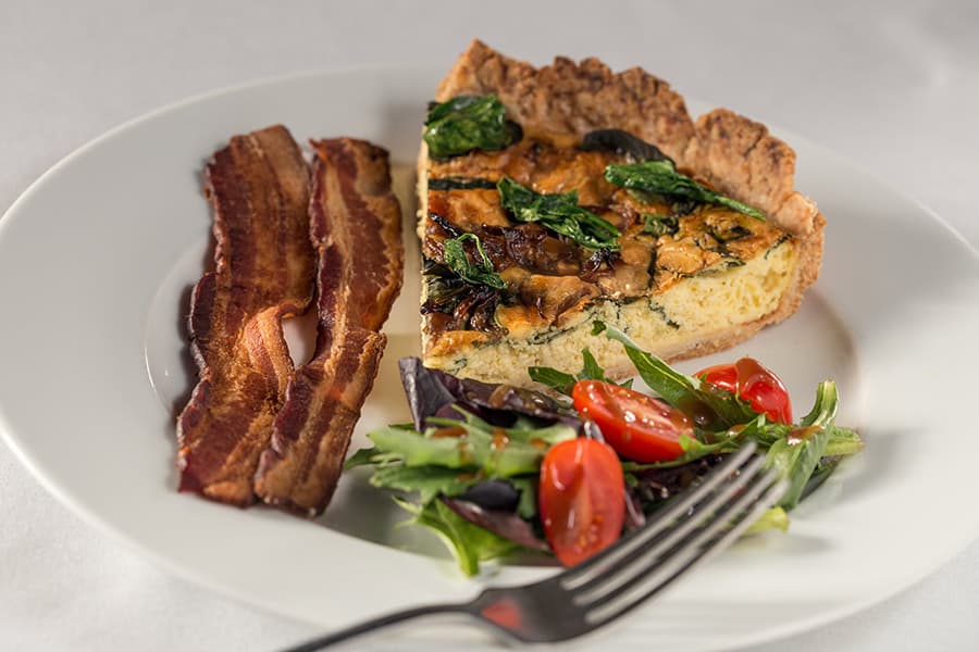 A plate of quiche with spinach, accompanied by crispy bacon and a fresh salad of mixed greens and cherry tomatoes.