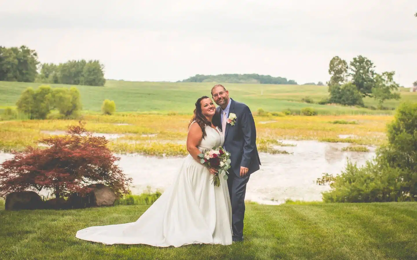 A joyful bride and groom pose together on a lush lawn with a scenic backdrop.