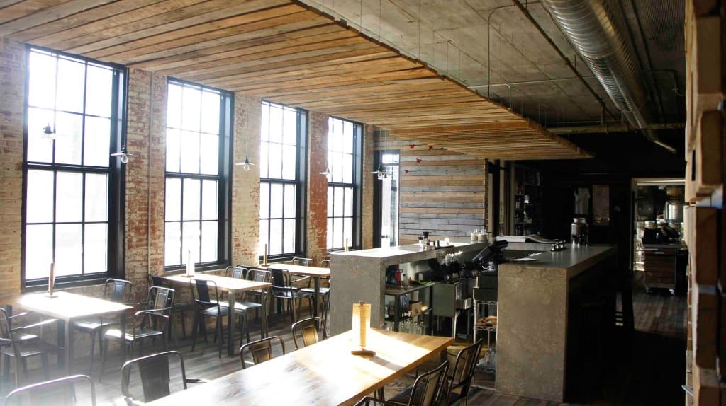 A spacious, industrial-style restaurant interior with large windows, wooden beams, and tables.