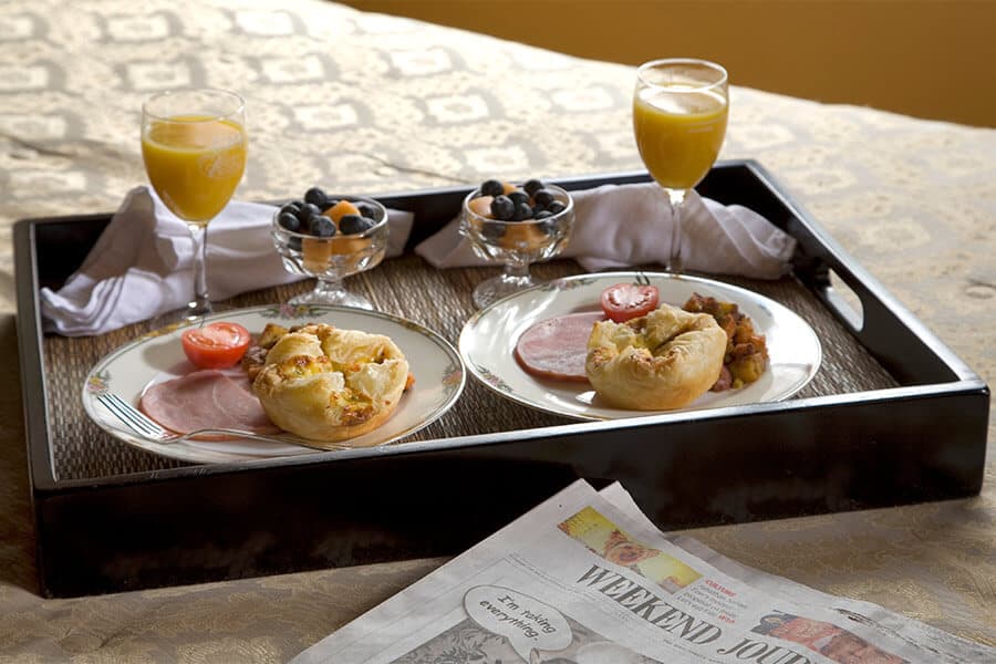 A breakfast tray featuring two plates with pastries, ham, and tomatoes, alongside fruit bowls and orange juice, resting on a decorative tablecloth with a newspaper.