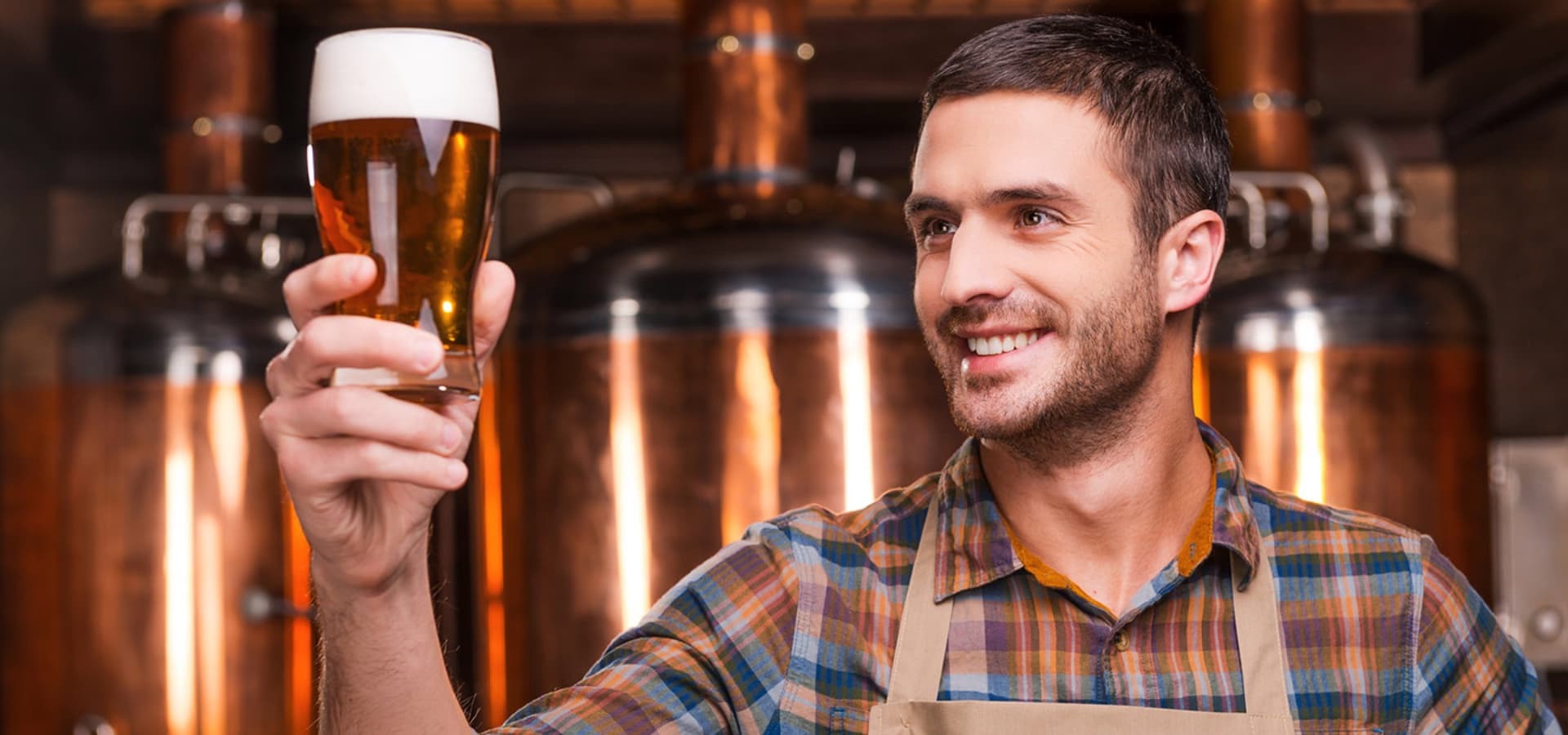 A smiling man in a plaid shirt holds up a glass of beer in a brewery setting.