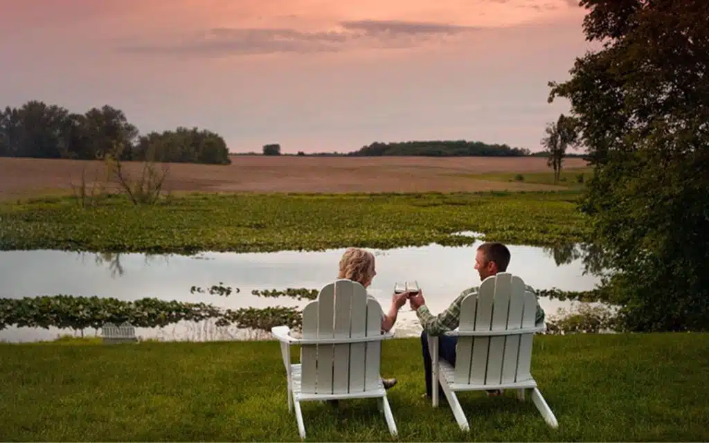 A couple toasts while sitting in white chairs by a tranquil pond at sunset.