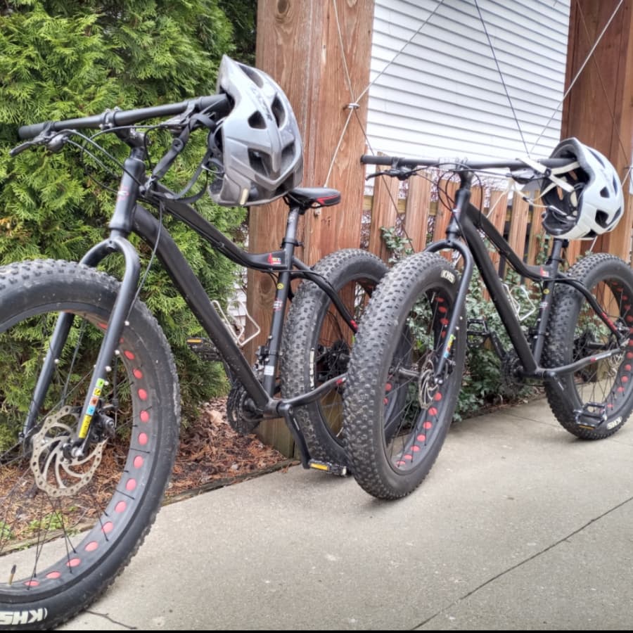Two black bicycles with fat tires and helmets are parked next to a wooden fence.