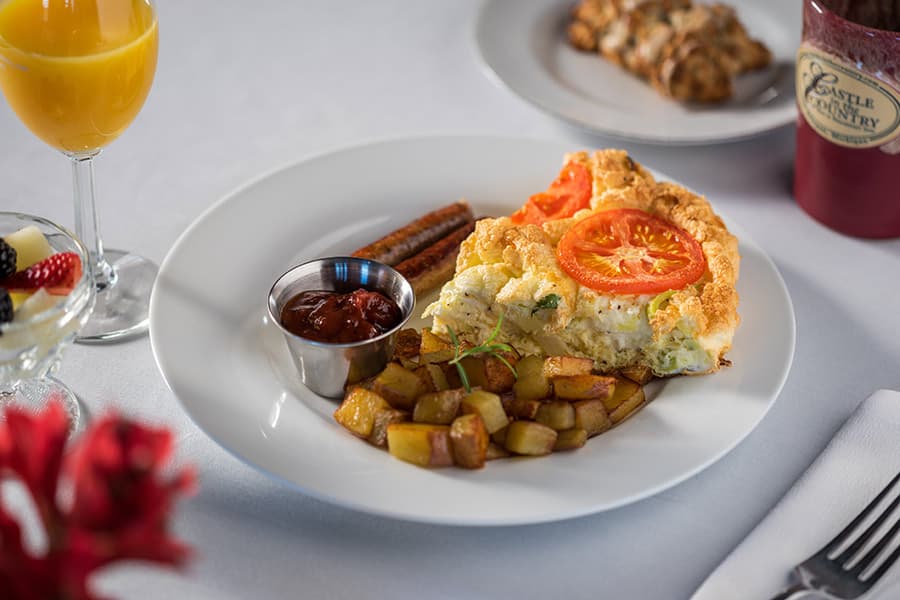 A plate of fluffy omelet topped with tomato, golden potatoes, sausage, and a side of salsa, accompanied by a mimosa and fruit.