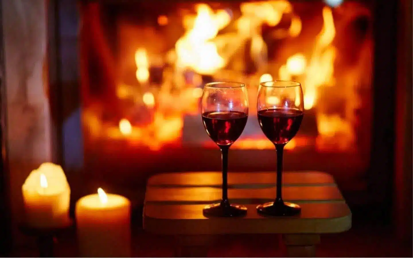 Two glasses of red wine on a table in front of a glowing fireplace, with candles nearby.