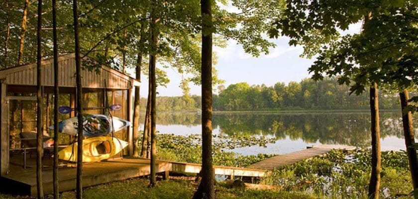 A serene lakeside view featuring a wooden cabin, a dock, and kayaks surrounded by lush greenery.