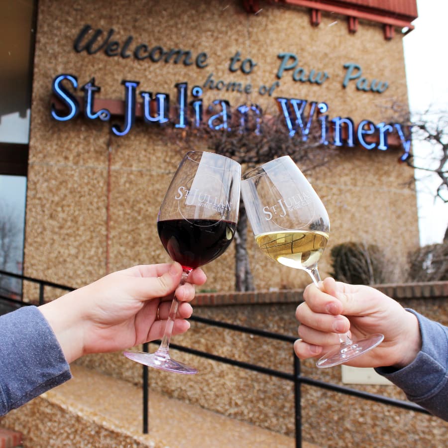 Two hands clink glasses of red and white wine in front of the St. Julian Winery sign.