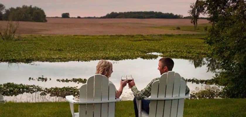 A couple sits in Adirondack chairs by a serene lake, toasting with drinks at sunset.