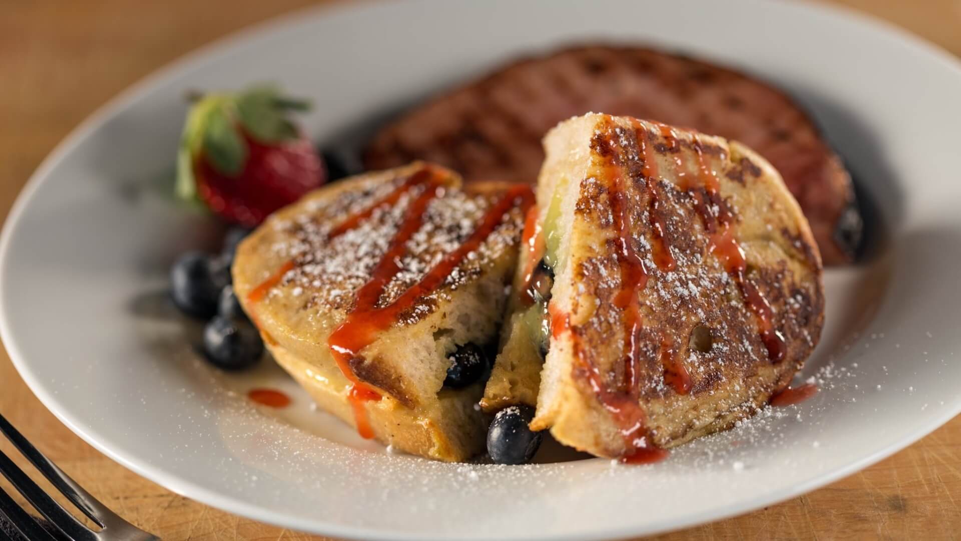 A plate of stuffed French toast with a strawberry, blueberries, and a slice of sausage.