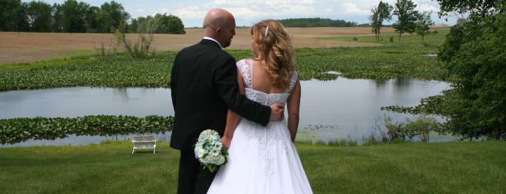 A couple in formal wedding attire stands together by a tranquil pond, looking out at the scenic landscape.