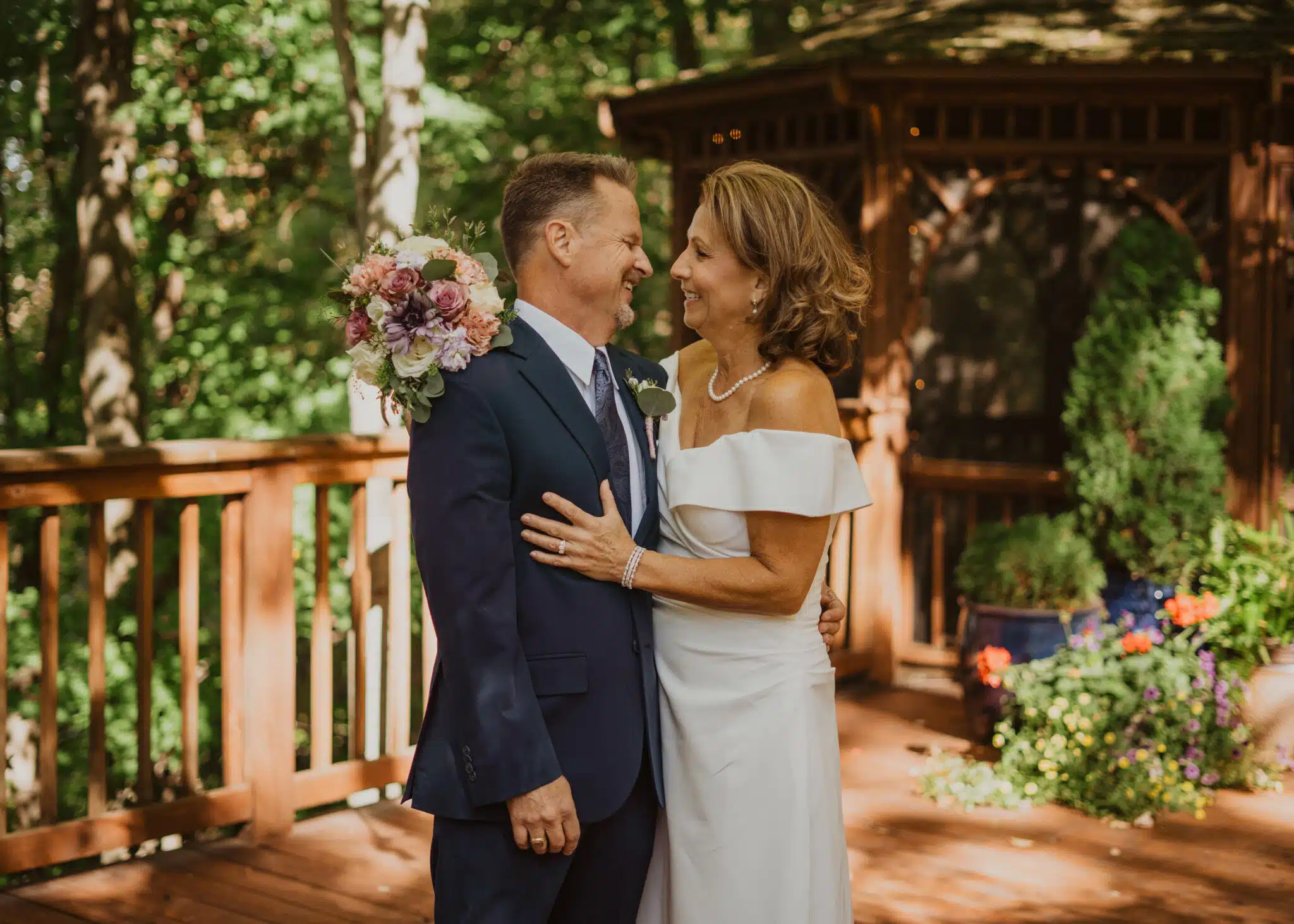 A couple joyfully embraces on a wooden deck surrounded by greenery, with one holding a bouquet.