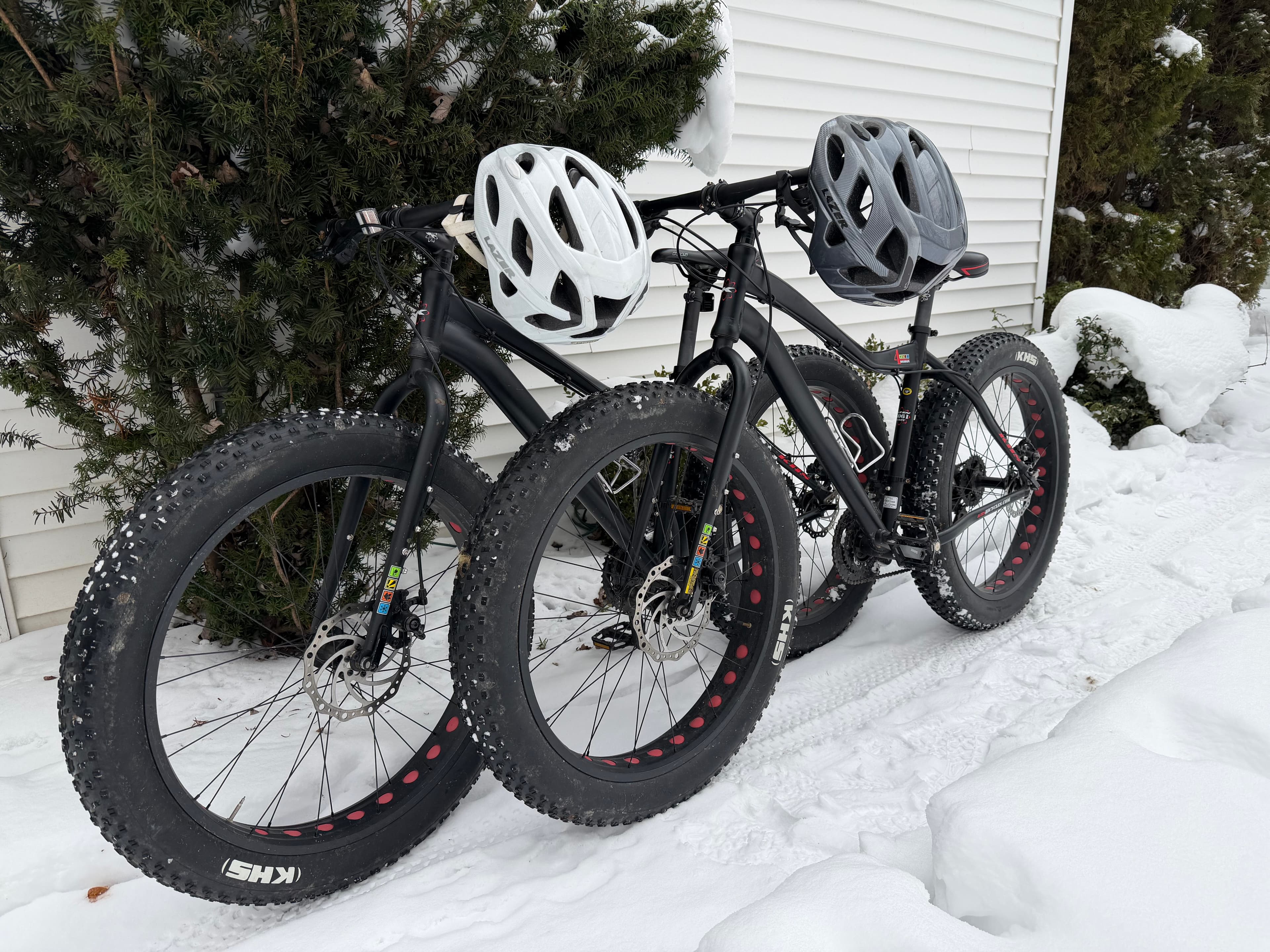 Two black fat bikes with helmets parked on snowy ground next to a house.