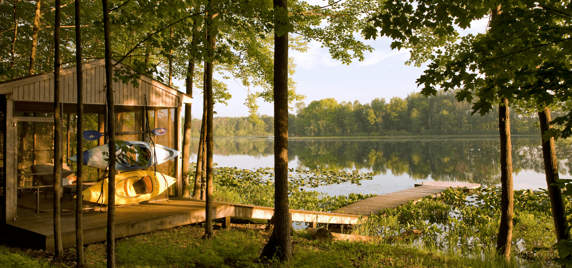 A serene lakeside scene featuring a wooden cabin with kayaks and a wooden dock surrounded by lush greenery.