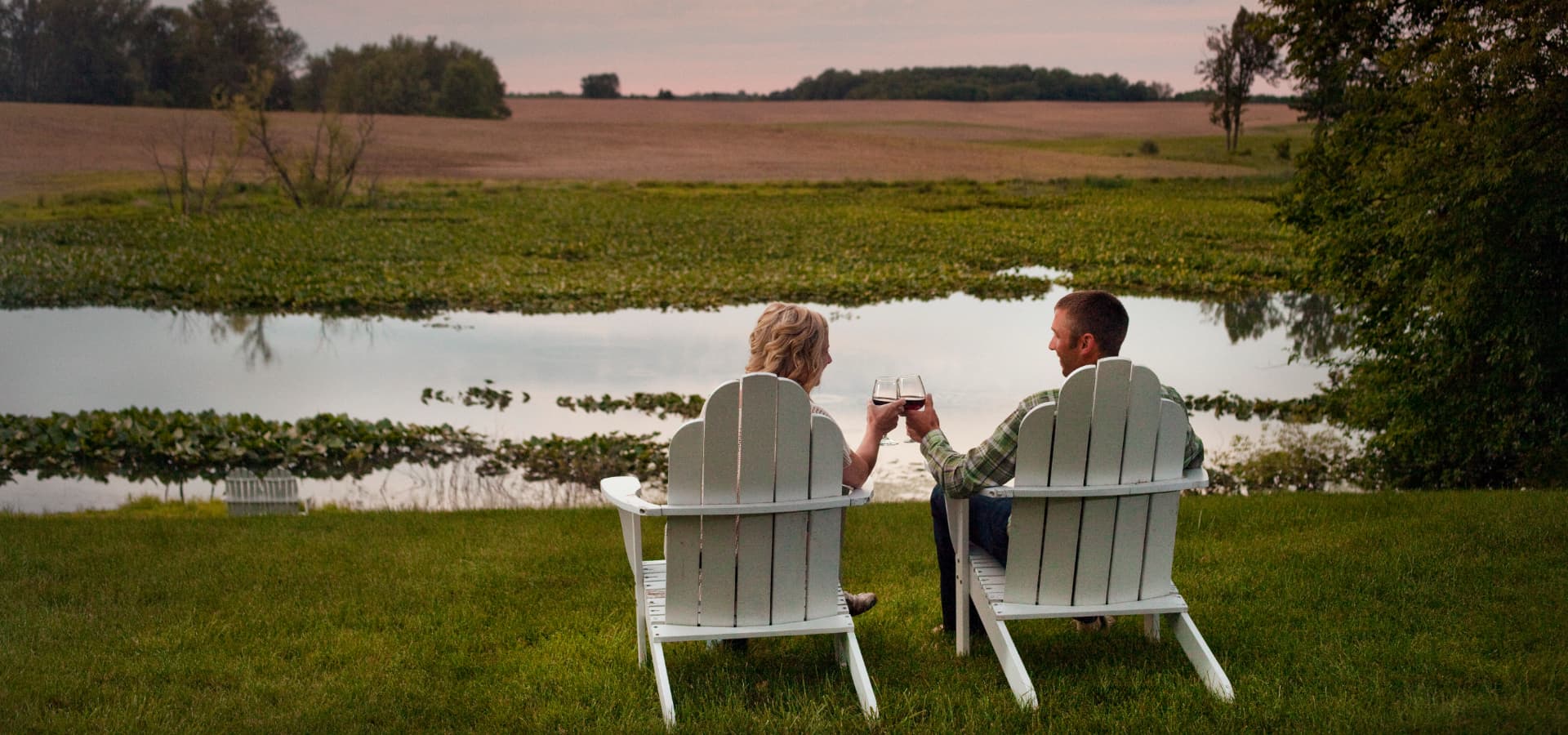 A couple clinks wine glasses while sitting in white chairs by a tranquil pond at sunset.