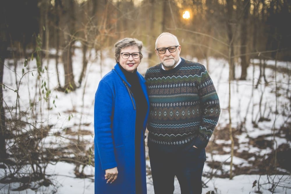 A couple stands together in a snowy forest at sunset, with the woman in a blue coat and the man in a patterned sweater.