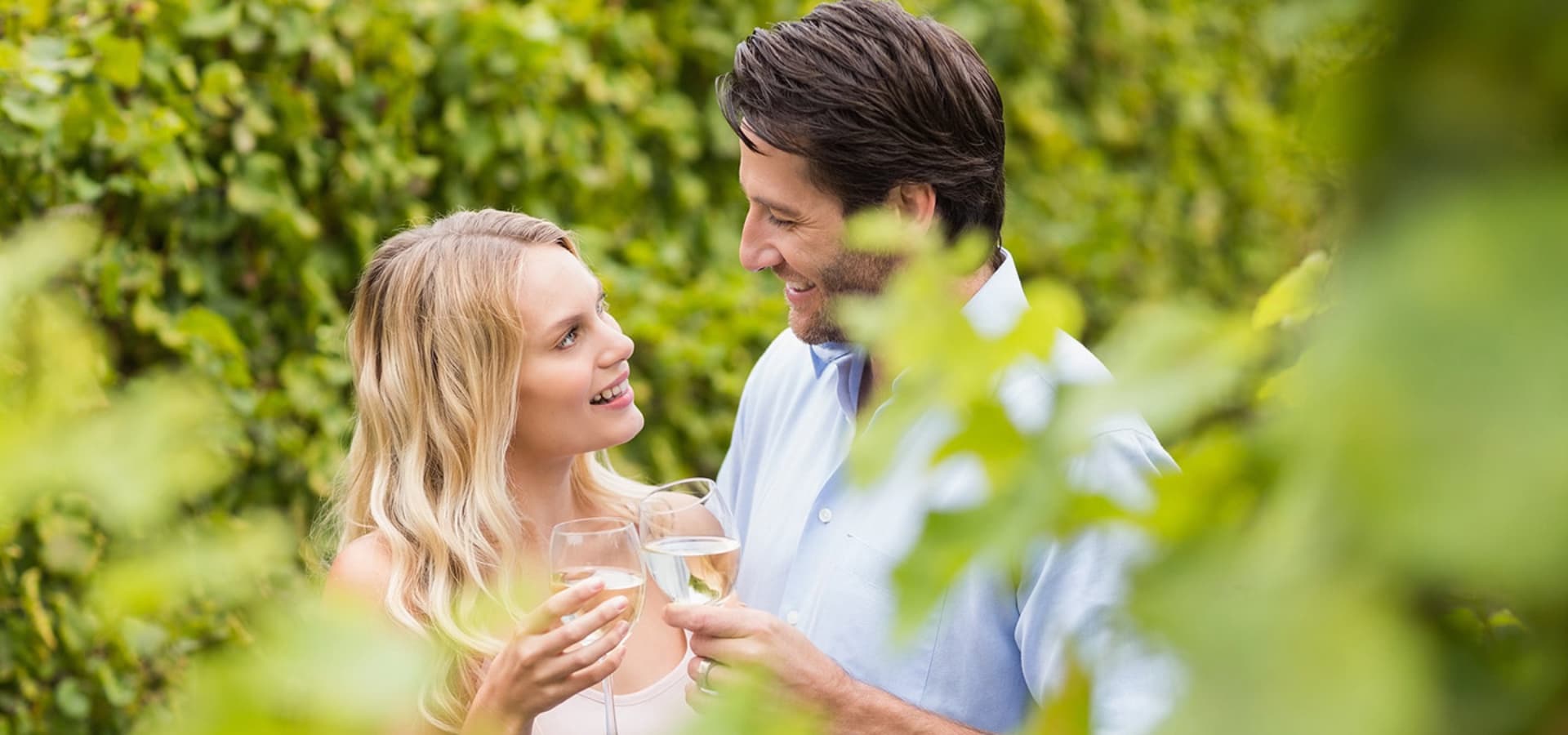 A couple smiles at each other while toasting with glasses of wine in a lush garden.