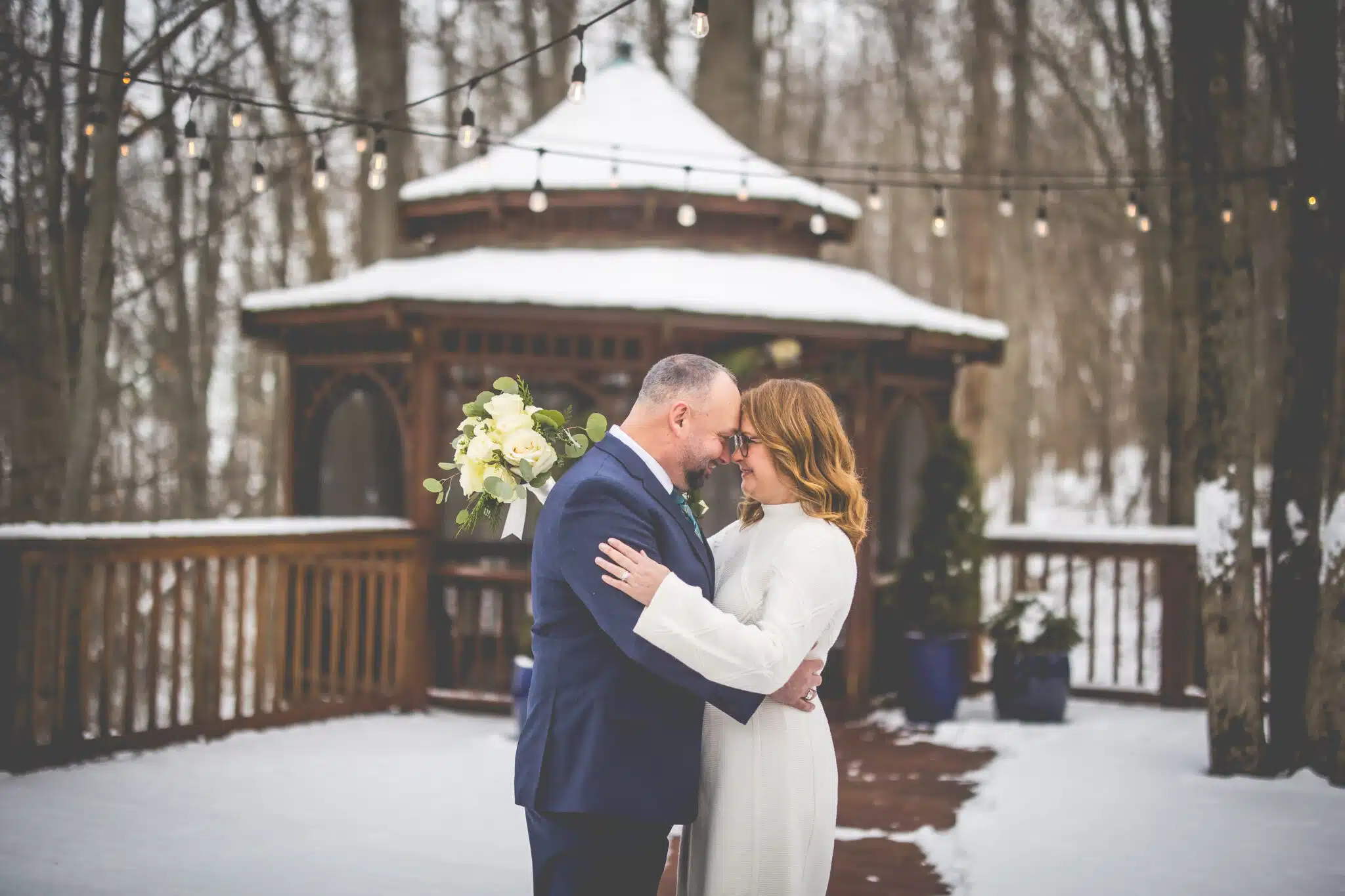 A couple embraces lovingly outdoors in the snow, surrounded by string lights and a gazebo.