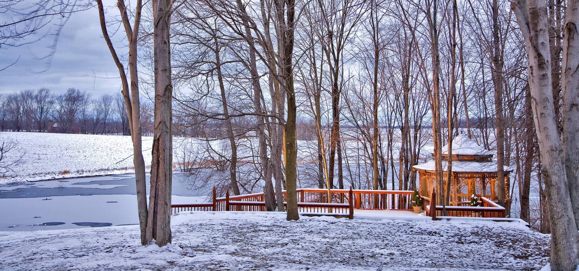 A snow-covered landscape featuring trees and a gazebo by a frozen lake.