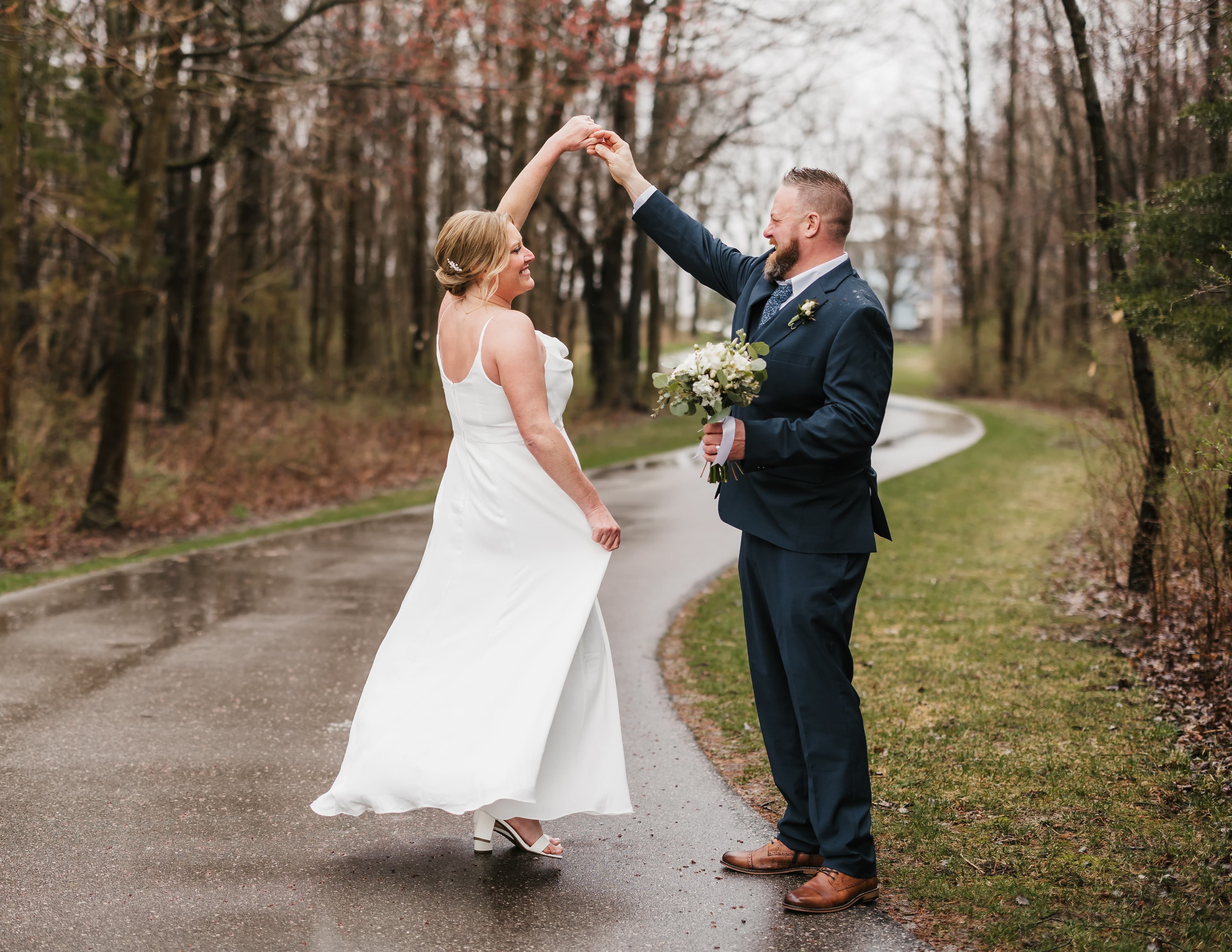 A bride in a white dress twirls with her groom in a dark suit along a rainy, tree-lined path.