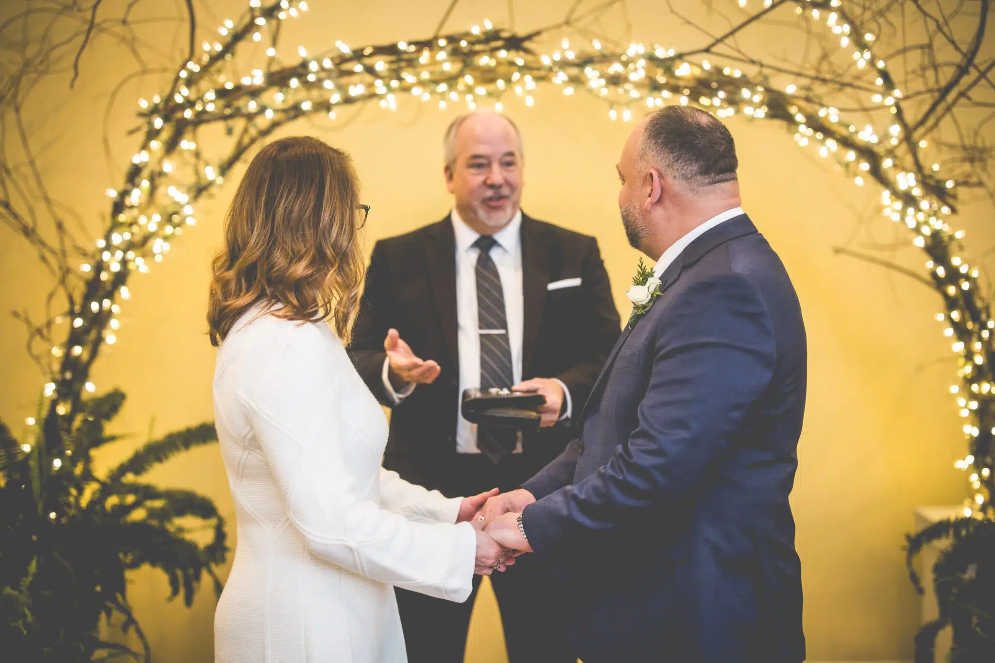 A couple stands hand-in-hand during their wedding ceremony officiated by a man against a backdrop of fairy lights.
