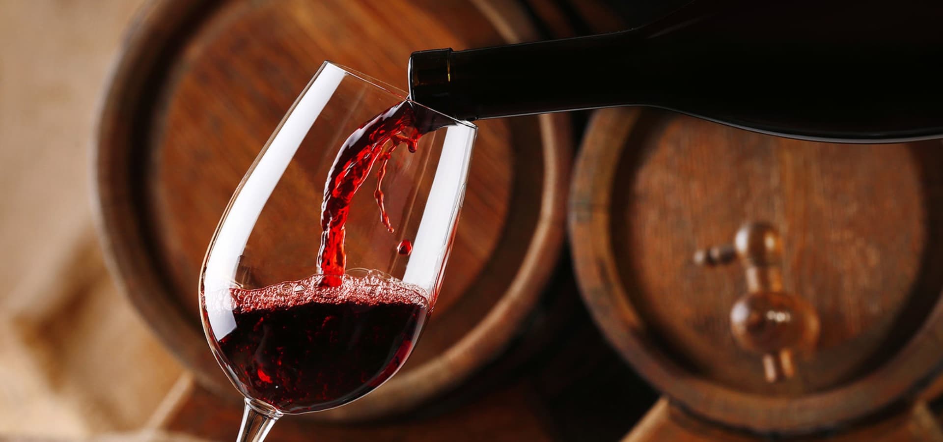 A red wine bottle pours wine into a glass against a backdrop of wooden barrels.