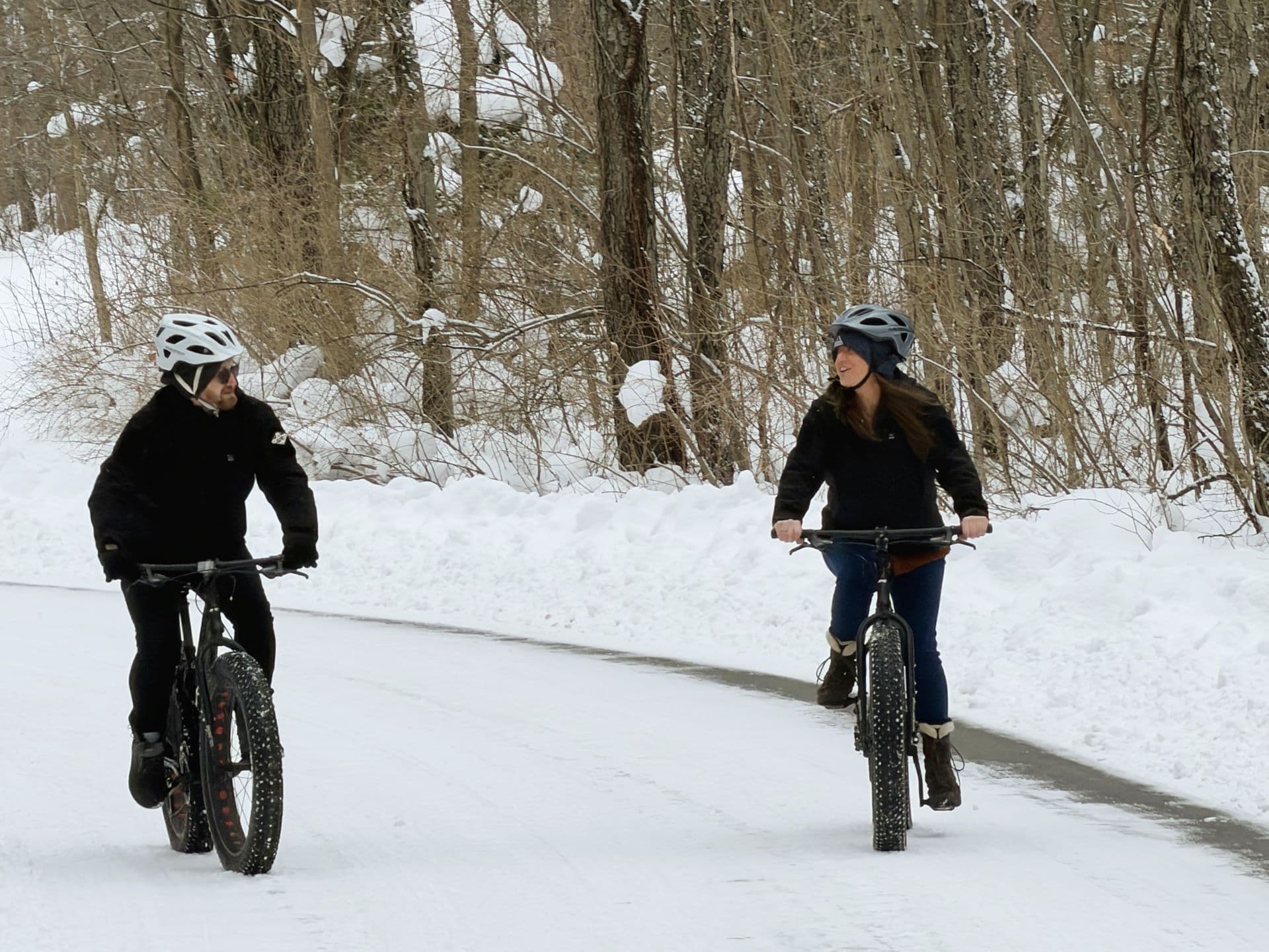 Two people ride fat bikes on a snow-covered path surrounded by trees.