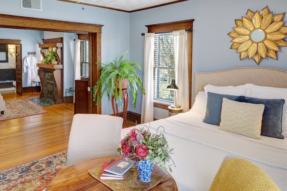 A cozy, well-decorated bedroom with a sunlit corner featuring a plant, a floral arrangement, and elegant furnishings.
