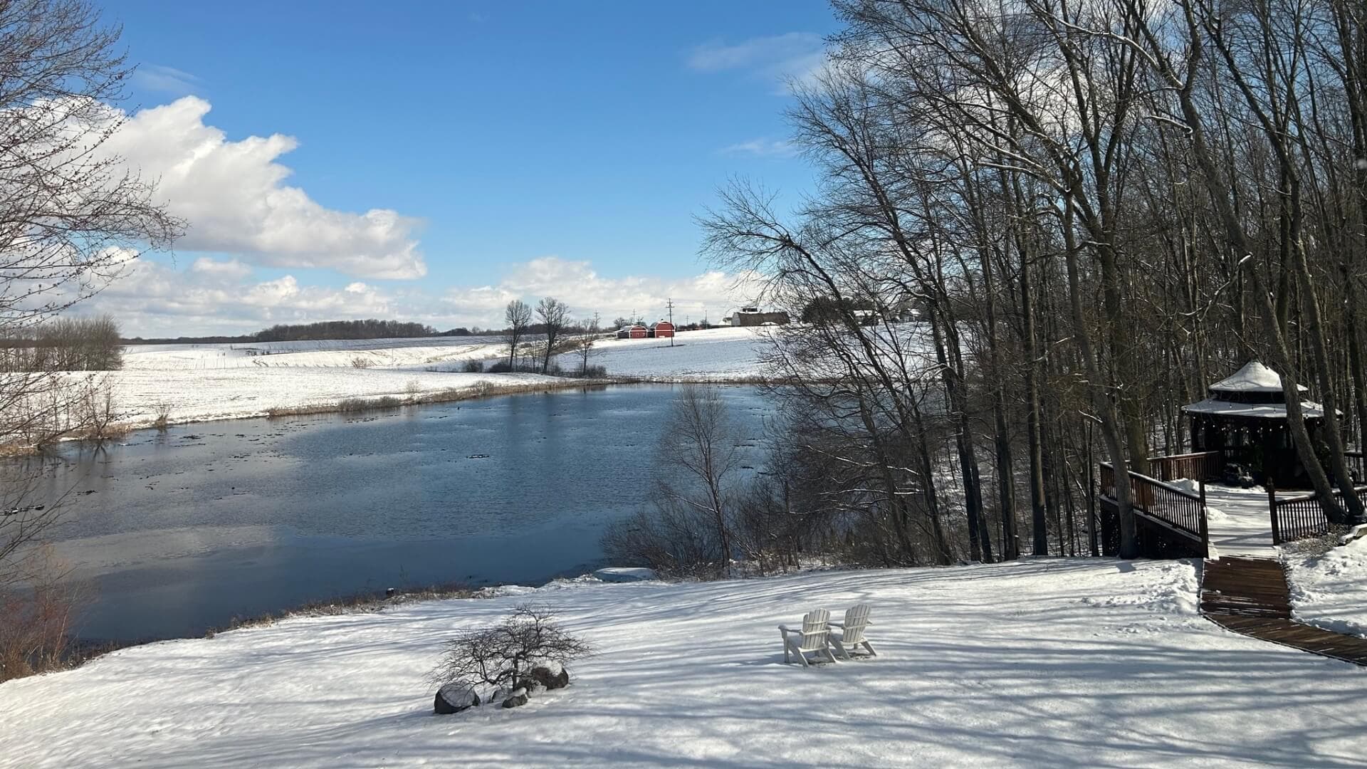 A snowy landscape with a river, barren trees, and a small dock overlooking a farm in the distance under a blue sky.